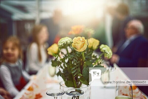 Vase mit frischen gelben und orangefarbenen Blumen als Tischdekoration bei einer Dinnerparty mit selektivem Fokus auf das Arrangement und unscharfen Personen