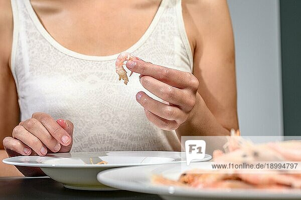 Frau  die an einem Esstisch sitzt und rosa Garnelen ißt oder zubereitet  während sie vorsichtig ihre Schalen für ein Gourmet Meeresfrüchteessen entfernt