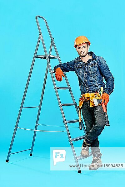 Ganzkörperporträt eines gut aussehenden männlichen Bauarbeiters  der auf einer Leiter über einem blaün Hintergrund klettert und zur Kamera schaut