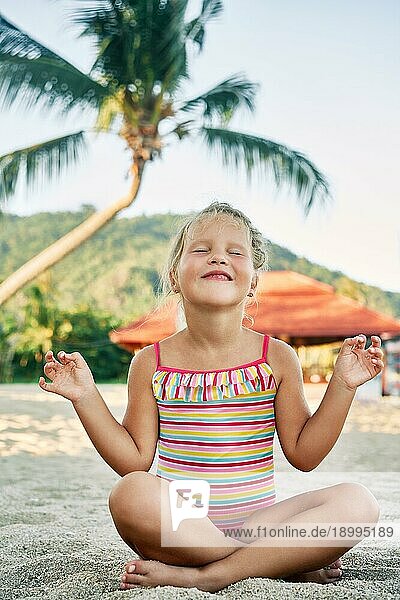 Hübsches kleines Mädchen meditiert in Lotuspose am Sandstrand im Sommerurlaub. Glückliche Kindheit Konzept