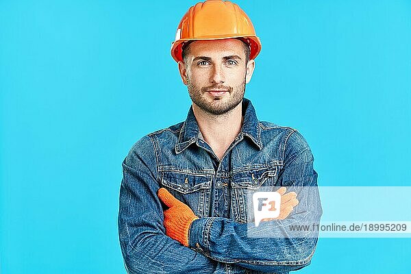 Porträt eines selbstbewussten  gut aussehenden männlichen Bauarbeiters mit Sicherheitshelm und verschränkten Armen  der auf einem blaün Hintergrund steht und zur Kamera schaut