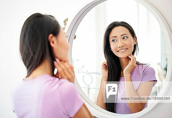Lächelnde schöne asiatische Frau  die sich zu Hause im Spiegel betrachtet. Spiegelbild  weibliches Schönheitskonzept