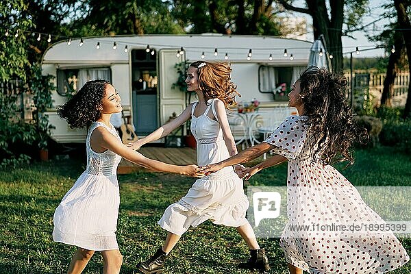 Glückliche schöne Frauen tanzen im Kreis während eines Picknicks. Multi ethnischen Mädchen Spaß haben zusammen im Freien. Kaukasische  afroamerikanische und asiatische Mädchen genießen den Urlaub. Frauen Freundschaft und Frauen Schönheit Konzept