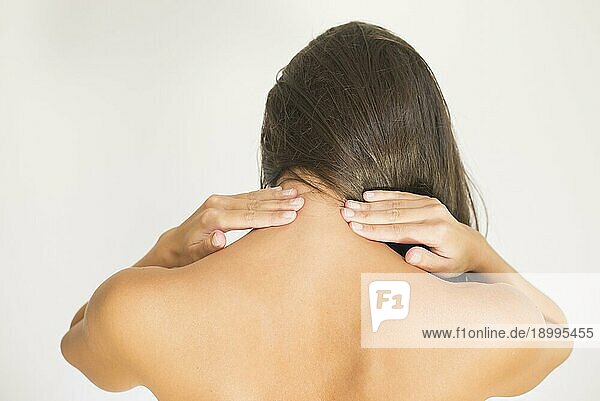 Frau mit Schmerzen im oberen Rücken und Nacken  die nackt mit dem Rücken zur Kamera steht und mit der Hand ihre Schultermuskeln nahe der Wirbelsäule reibt