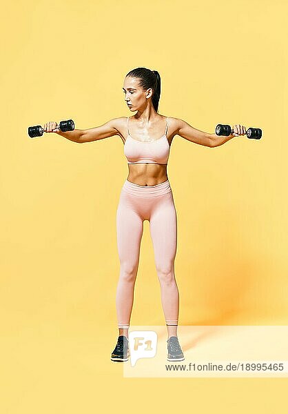 Sportliche Frau in Sportkleidung beim Training mit Hanteln auf gelbem Hintergrund. Konzept für Stärke und Motivation. Porträt in voller Länge