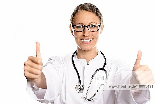 Motivierte attraktive Ärztin oder Krankenschwester im weißen Kittel und mit Stethoskop  die mit beiden Händen den Daumen nach oben streckt und damit Erfolg und eine positive Prognose und Diagnose anzeigt  vor weißem Hintergrund