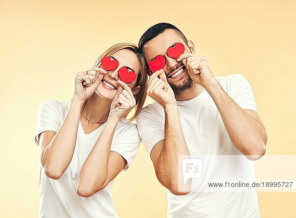 Happy couple have fun holding small paper hearts by their eyes over studio background. Liebe  Beziehung und romantisches Konzept
