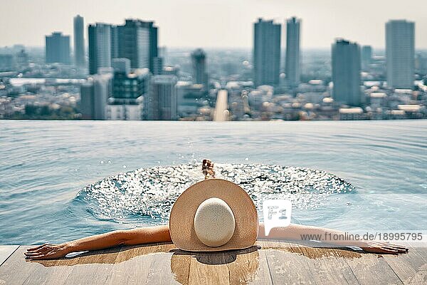 Rückansicht einer jungen Frau mit Hut  die sich im Schwimmbad auf dem Dach eines Hotels entspannt und das Stadtbild genießt. Sommerurlaub  Erholung Konzept