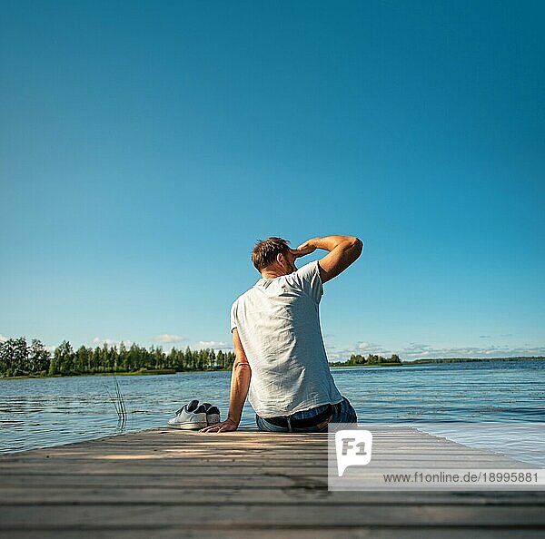 Mann sitzt auf einem Holzsteg am See im Sommer in Finnland