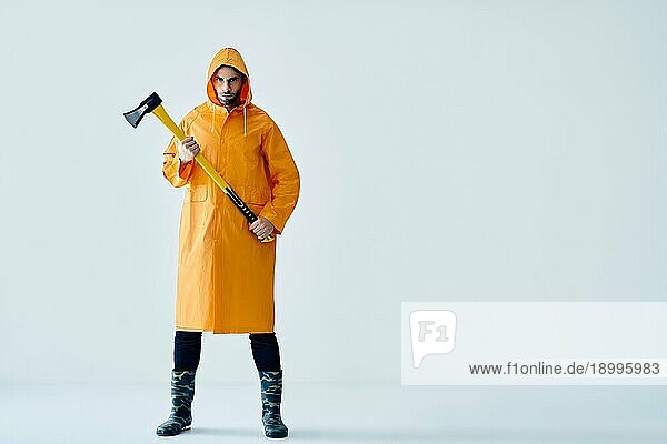 Ganzkörperporträt eines jungen  gut aussehenden Mannes in einem hellen Regenmantel  der eine große Axt hält  mit Kopierraum. Brutaler Männchen  Holzfäller  Fischer mit Axt