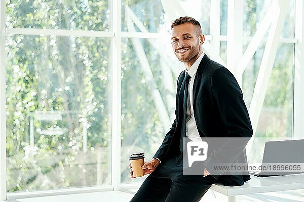Glücklich gut aussehend Geschäftsmann entspannen mit einer Tasse Kaffee in der Hand während der Pause in einem modernen Büro mit Kopie Raum. Ruhe  Erfolg Konzept