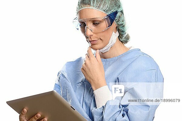 Attraktive weibliche Chirurgin oder Arzt in einer Maske Schutzbrille und chirurgischen Kittel stehend Beratung ein Tabletcomputer lesen die Informationen mit einem ernsten Ausdruck  vor weißem Hintergrund