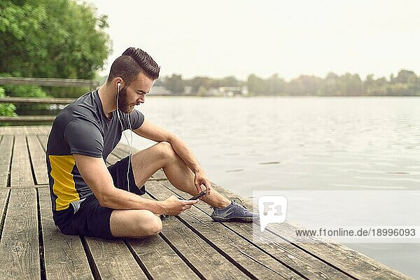 Attraktiver  bärtiger junger Mann  der sich auf einer Holzterrasse mit Blick auf einen ruhigen See entspannt und mit seinem Handy in der Hand lächelnd in die Kamera schaut