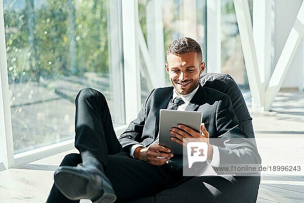 Lächelnde erfolgreiche Geschäftsmann in eleganten Suite mit Tablet sitzen auf Sitzsack Stuhl mit Panoramafenstern Hintergrund in modernen kreativen Büro