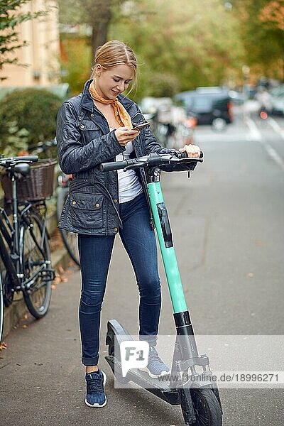 Junge Frau mit elektrischen Kick Scooter in der Straße der Stadt  Blick auf ihr Telefon und lächelnd. Öffentliche Verkehrsmittel Vermietung Service  moderne städtische ökologische Null Emissions Transport Konzept