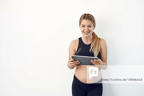 Glückliche junge schwangere Frau mit nacktem Babybauch  die mit einem zufriedenen Lächeln auf einem Tablet PC liest  vor einem weißen Studiohintergrund mit Kopierraum