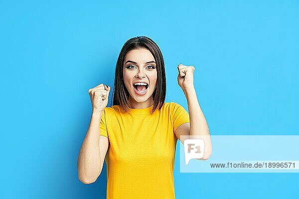 Glücklich aufgeregt junge Frau feiern Erfolg mit den Händen nach oben vorblauem Hintergrund. Sieg  Emotionen Konzept