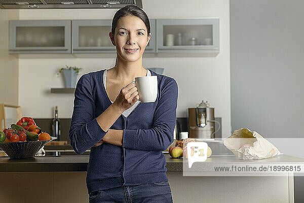 Glückliche  zufriedene Hausfrau in ihrer Küche  die der Kamera ein warmes  freundliches Lächeln schenkt  während sie sich mit einer Tasse Kaffee und frischem Obst auf dem Tresen hinter ihr entspannt