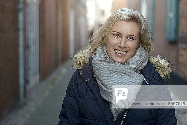 Attraktive  freundliche blonde Frau mit modischem Winterschal  die in einer engen Gasse in der Stadt steht und fröhlich in die Kamera lächelt