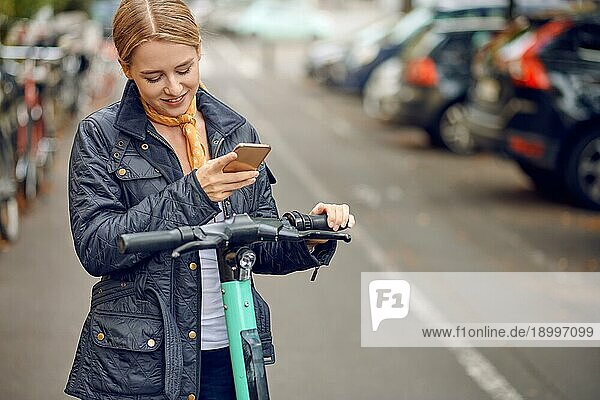 Glückliche junge Frau in lässiger grauer Jacke  die in die Kamera schaut und lächelt  steht auf der Straße mit einem escooter und einem Handy in der Hand