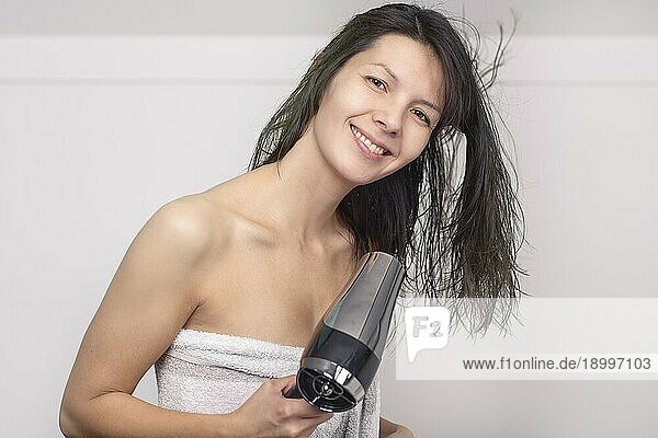 Attraktive Frau in einem Handtuch föhnt ihr braunes Haar mit einem Handfön in ihrem Badezimmer und schaut mit einem warmen  freundlichen Lächeln in die Kamera
