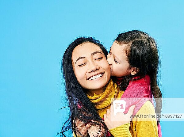 Kleine süße Tochter umarmen und küssen ihre glückliche Mutter vorblauem Hintergrund. Familie Liebe Konzept