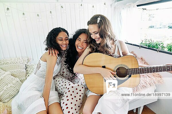 Attraktive glückliche Frauen verschiedener Ethnien mit Gitarre haben Spaß zusammen im Inneren des Wohnmobils. Frauen Freundschaft und Frauen Schönheit Konzept. Kaukasisch  afrikanisch amerikanischen und asiatischen Mädchen umarmen und genießen Sie zu Hause Partei