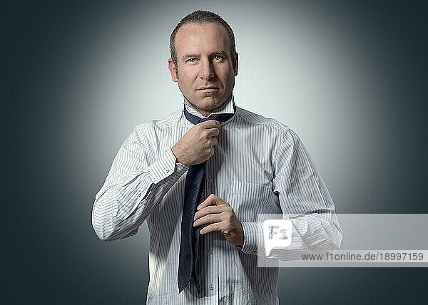 Halbkörperaufnahme eines erwachsenen Geschäftsmannes  der eine Krawatte bindet und in die Kamera lächelt  vor grauem Hintergrund