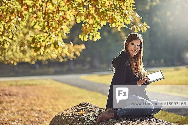 Hübsche junge Frau entspannt sich in einem herbstlichen Park  liest auf ihrem Tablet PC und lächelt fröhlich in die Kamera