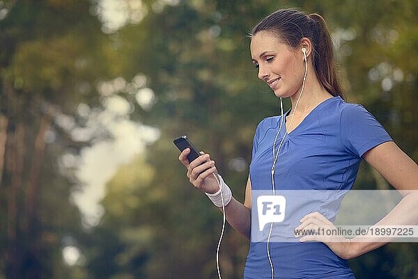 Attraktive  glückliche Frau  die mit ihrem MP3 Player und Ohrstöpseln auf einem unbefestigten Waldweg steht und mit einem Lächeln der Freude im Gesicht Musik hört