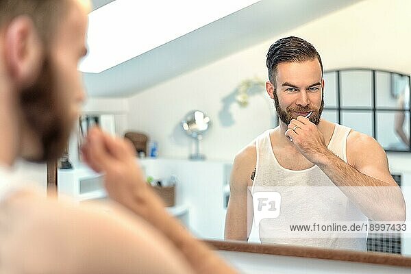 Attraktiver junger bärtiger Mann  der sich im Badezimmer die Zähne putzt und sich dabei im Spiegel betrachtet  in einem Zahnpflegekonzept