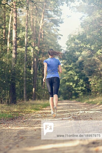 Sportliche junge Frau in Sportkleidung  die auf einem Waldweg im Rahmen eines Fitness und Wellnesskonzepts von der Kamera weg joggt