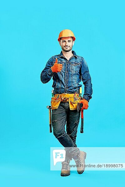 Porträt eines selbstbewussten  gut aussehenden männlichen Bauarbeiters mit Schutzhelm  der ein Daumen hoch Zeichen vor blauem Hintergrund zeigt und in die Kamera blickt  in voller Länge