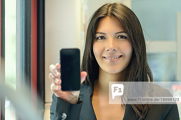 Attraktive Frau  die ihr Mobiltelefon mit dem leeren Bildschirm zum Betrachter hochhält  während sie mit einem glücklichen  warmen  freundlichen Lächeln in die Kamera blickt  drinnen zu Hause mit Fokus auf ihr Gesicht