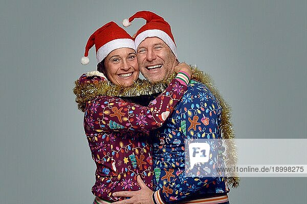 Ein fröhliches Ehepaar feiert Weihnachten zusammen in festlichen roten Weihnachtsmannmützen und Weihnachtsmänteln  die sich umarmen und lachen  während sie in die Kamera lächeln