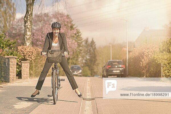 Elegante Frau in einem lockeren Anzug und mit Schutzhelm  die Spaß daran hat  auf ihrem Fahrrad zur Arbeit zu fahren  indem sie ihre Füße in die Luft hebt und balanciert  während sie mit einem glücklichen Lächeln eine Wohnstraße entlangfährt