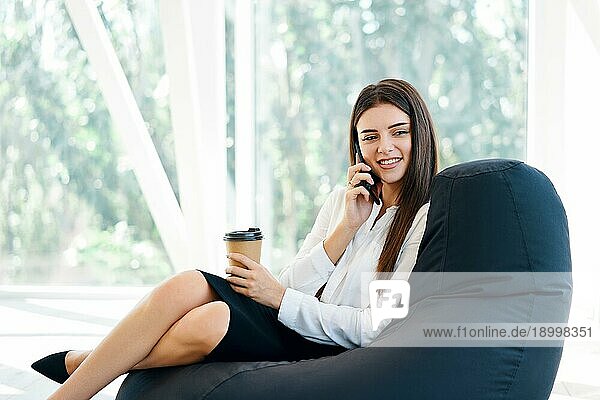 Hübsche Geschäftsfrau spricht am Telefon sitzen auf Sitzsack Stuhl hält Tasse Kaffee in modernen kreativen Büro mit Panoramafenster Hintergrund i. Pause Zeit Konzept  geschäftlichen Erfolg