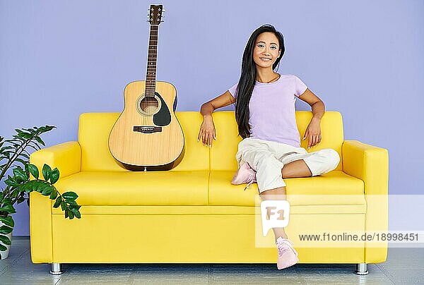 Hübsche Frau mit Gitarre sitzt auf einem bequemen Sofa und schaut in die Kamera
