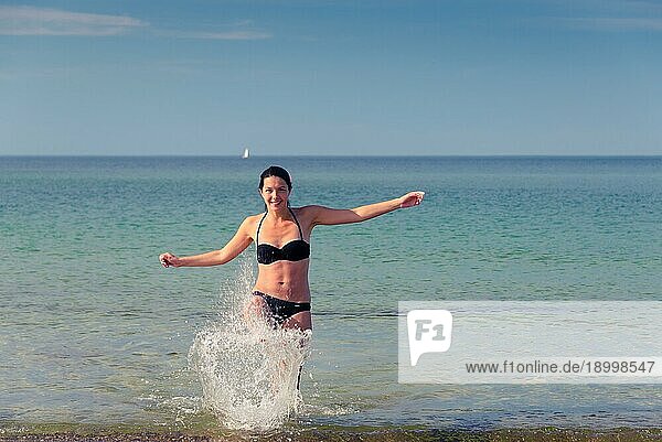 Fröhliche attraktive junge Frau im Bikini  die an einem tropischen Strand herumtollt  lacht und mit dem Fuß Gischt aus dem Meer aufwirbelt  mit Kopierraum nach rechts
