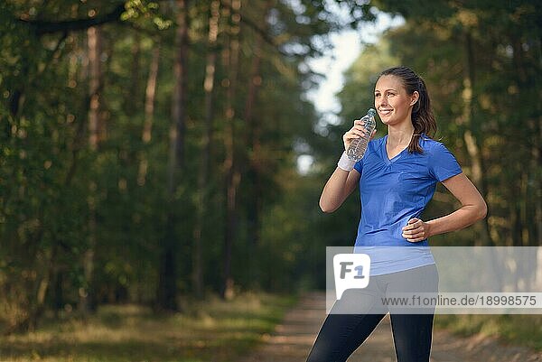 Fitte schlanke junge Frau in Sportkleidung trinkt Wasser in Flaschen  während sie auf einem Waldweg eine Pause einlegt  um während eines Trainingslaufs auf dem Lande neue Flüssigkeit zu sich zu nehmen  Gesundheits und Fitnesskonzept