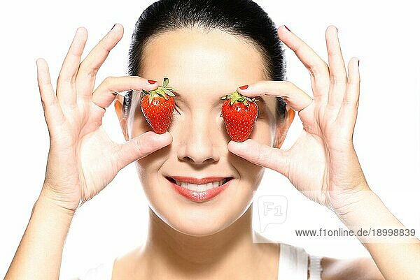 Schöne junge Frau mit einem schönen warmen Lächeln und gesunder gebräunter Haut  die zwei reife rote Erdbeeren an die Seiten ihrer Augen hält  vor weißem Hintergrund