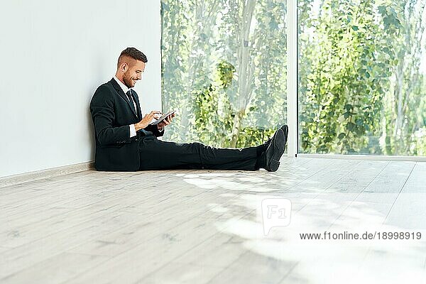Junge lächelnde Geschäftsmann sitzt auf dem Boden in seinem Büro und Blick auf Smartphone. Pause Zeit Konzept  Business Technologie