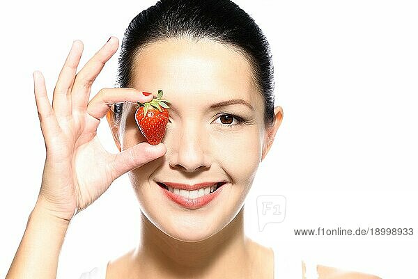Schöne verführerische gebräunte junge Frau mit einer reifen Erdbeere  die sie an ihr Auge hält und ihre Lippen in einer sinnlichen  provokativen Weise aufreißt  während sie in die Kamera schaut  vor weißem Hintergrund