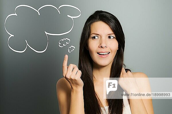 Attraktive junge Frau signalisiert mit ihrem Zeigefinger eine Idee in Form einer illustrierten Sprechblase  auf einem grauen Studiohintergrund