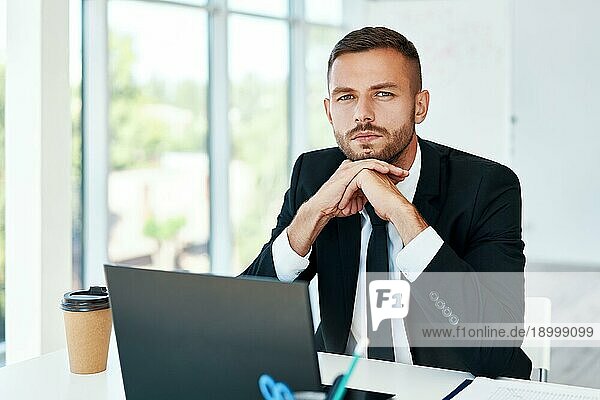 Selbstbewusster junger Geschäftsmann in elegantem Anzug posiert in einem hellen  modernen Büro. sitzt an seinem Schreibtisch. Business Erfolg Konzept