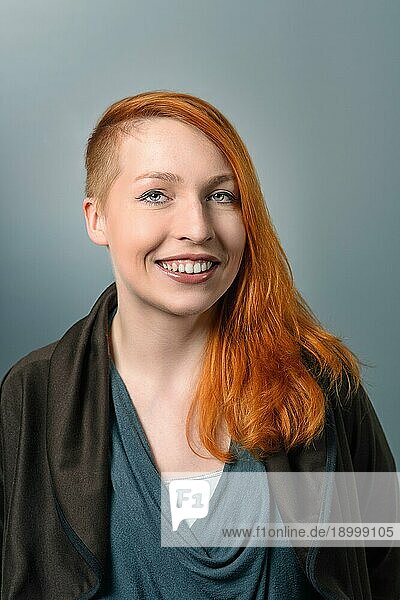 Porträt einer lächelnden  zuversichtlichen rothaarigen Frau mit einer seitlich geschnittenen Frisur  die in die Kamera schaut  auf grauem Studiohintergrund