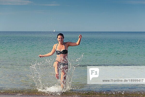 Fröhliche attraktive junge Frau im Bikini  die an einem tropischen Strand herumtollt  lacht und mit dem Fuß Gischt aus dem Meer aufwirbelt  mit Kopierraum nach rechts
