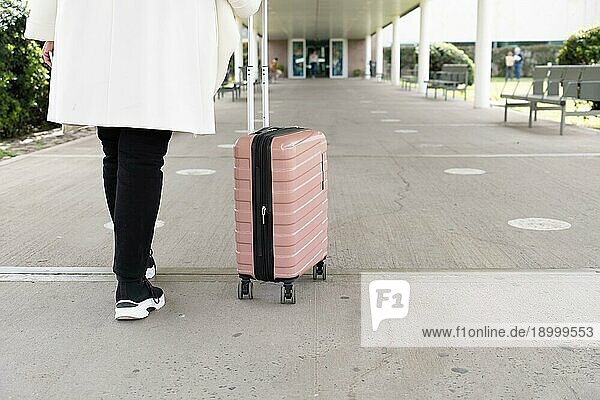 Aufnahme der Beine einer Frau in Turnschuhen und Mantel  die zum Haupteingang des Flughafens geht und ihren Rollkoffer rollt. Reisekonzept