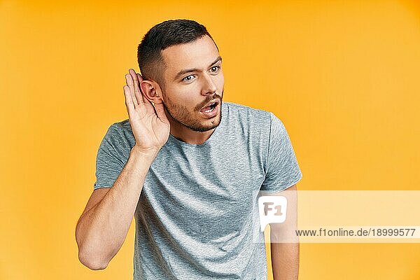 Junger überraschter Mann hört etwas aufmerksam zu und hält seine Hand am Ohr auf gelbem Hintergrund