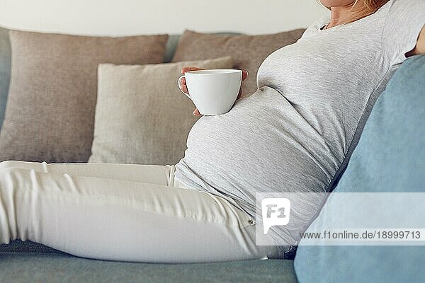 Schwangere Frau entspannt sich auf einem Sofa mit einem großen Becher mit heißem Getränk in einer Nahaufnahme Seitenansicht ihres Babybauchs und Hände halten die Tasse